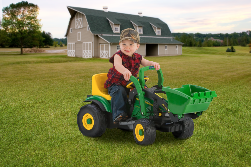 Toddler seated on green and yellow ride-on loader with front bucket on grass near large barn