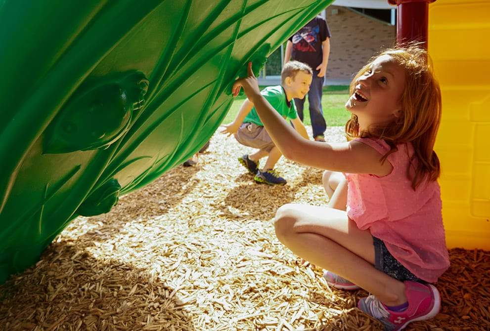 Children playing under a green climbing structure with textured grips and a yellow playhouse wall on wood chip ground