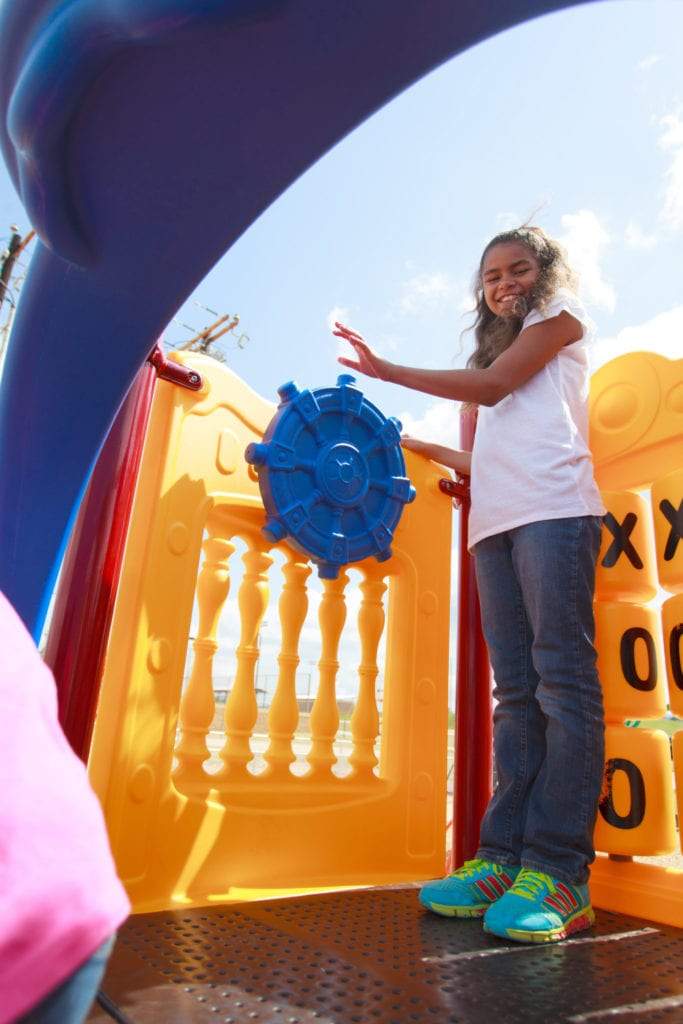 Child playing on colorful playground structure with blue ship’s wheel and tic-tac-toe panel under clear sky