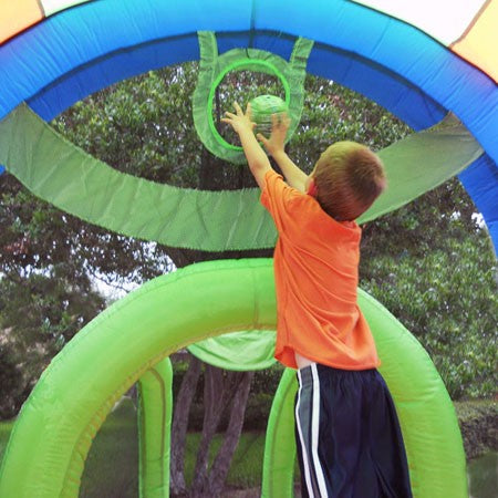 Child in orange shirt playing basketball inside inflatable sports bounce house with green and blue hoops outdoors
