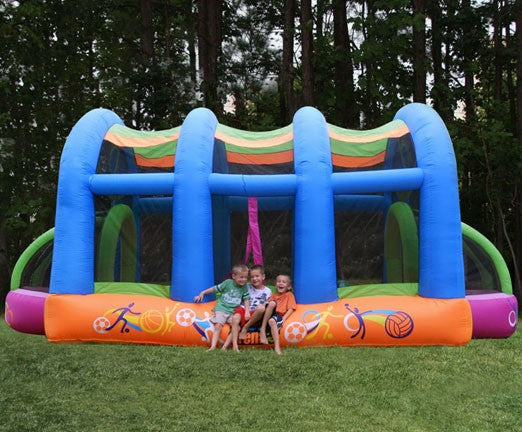 Colorful inflatable sports bounce house with arches, soccer goals, basketball hoops, and three children sitting on grass in front