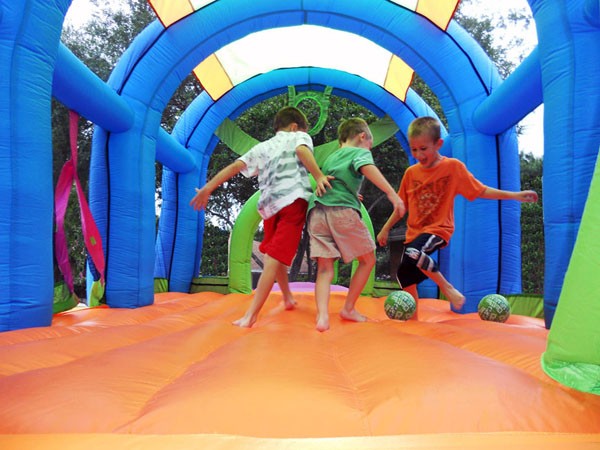 Three children playing soccer with inflatable balls inside a colorful blue and orange inflatable sports bounce house with goals and basketball hoops