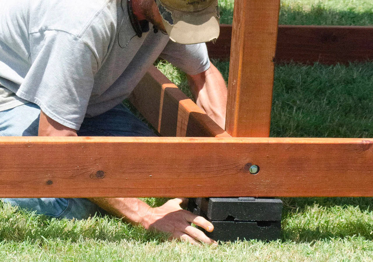 Man placing black plastic leveling blocks under wooden playset legs on grass to prevent moisture damage and stabilize structure
