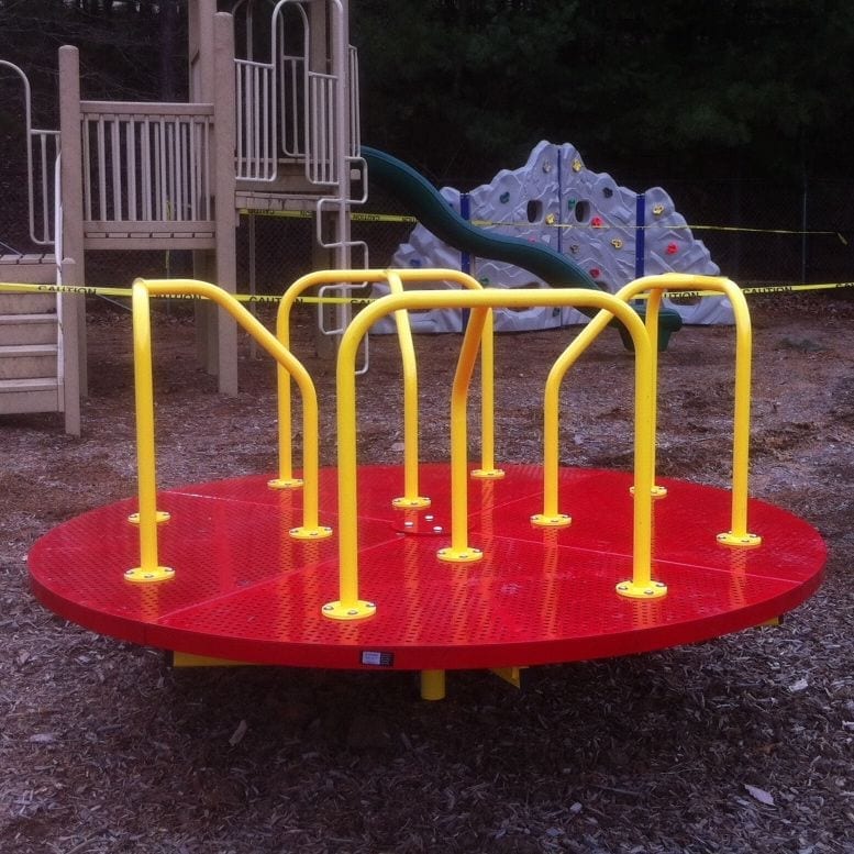 Red and yellow powder-coated steel merry go round with safety handrails on playground mulch near climbing wall and slide
