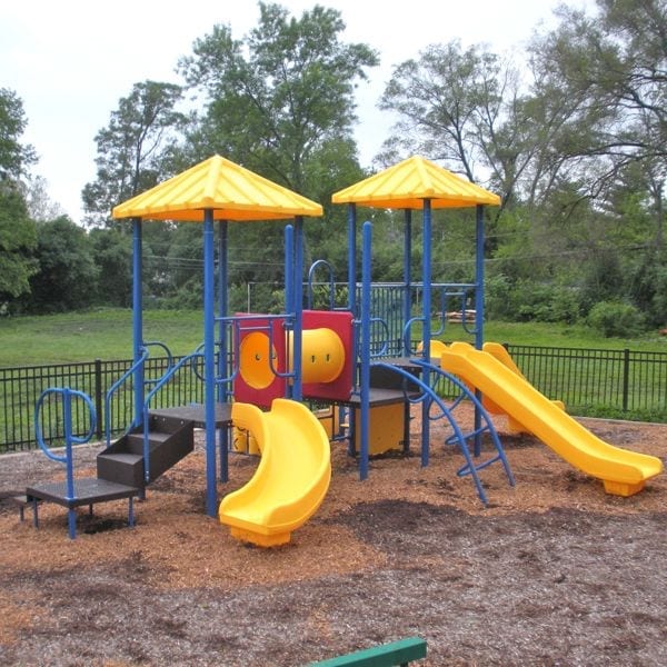 Outdoor modular playground with yellow slides, blue climbing structures, tic tac toe panel, and two yellow roofs on wood chip ground.