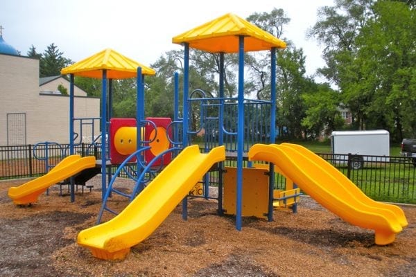 Yellow and blue modular playground with double and 90-degree slides, climbing structures, tic tac toe panel, and platforms on wood chips.