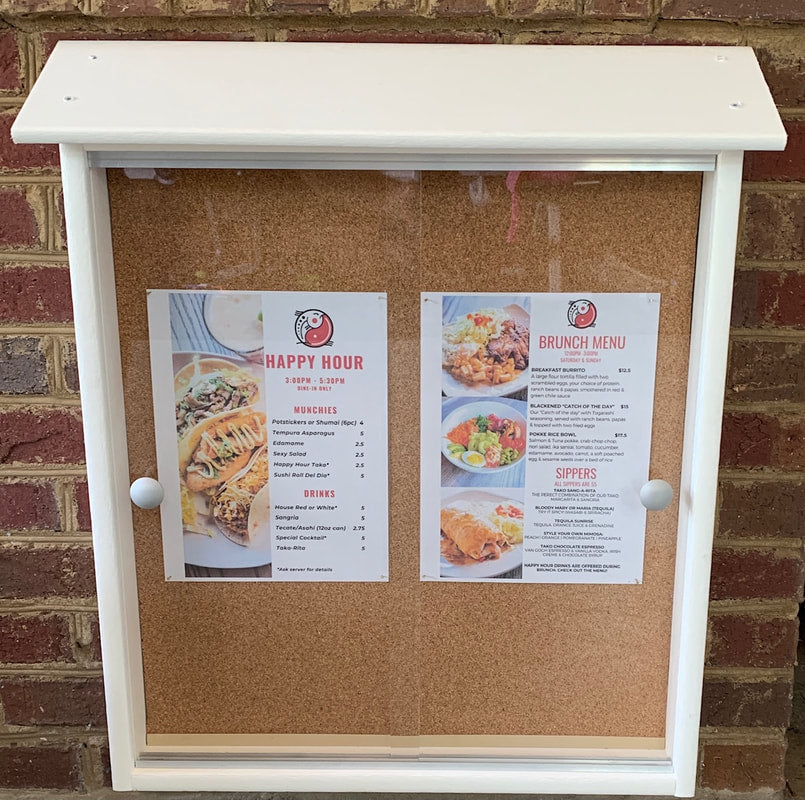 Outdoor white-framed cork bulletin board with sliding acrylic doors displaying two menus featuring food and drink items