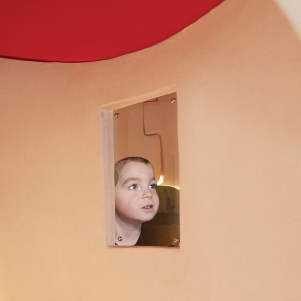Child peeking through a small window of a whimsical red-roofed mushroom-shaped playhouse with tactile shapes and a side bench