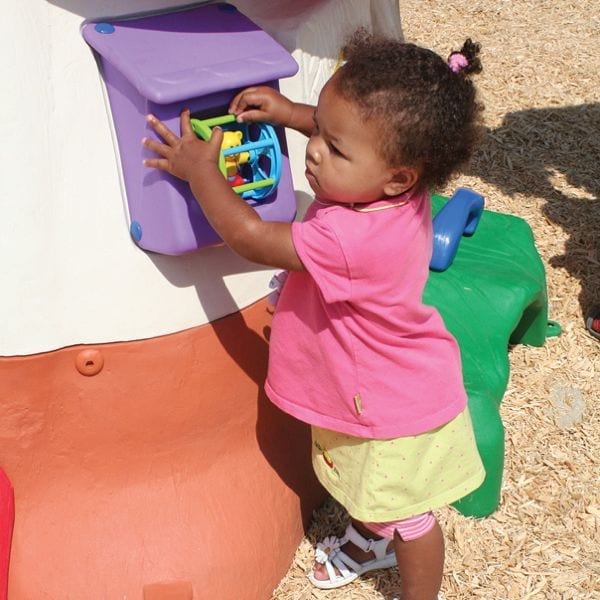 Toddler engaging with colorful manipulative play panel on mushroom-shaped outdoor playhouse with green bench and toadstool chair