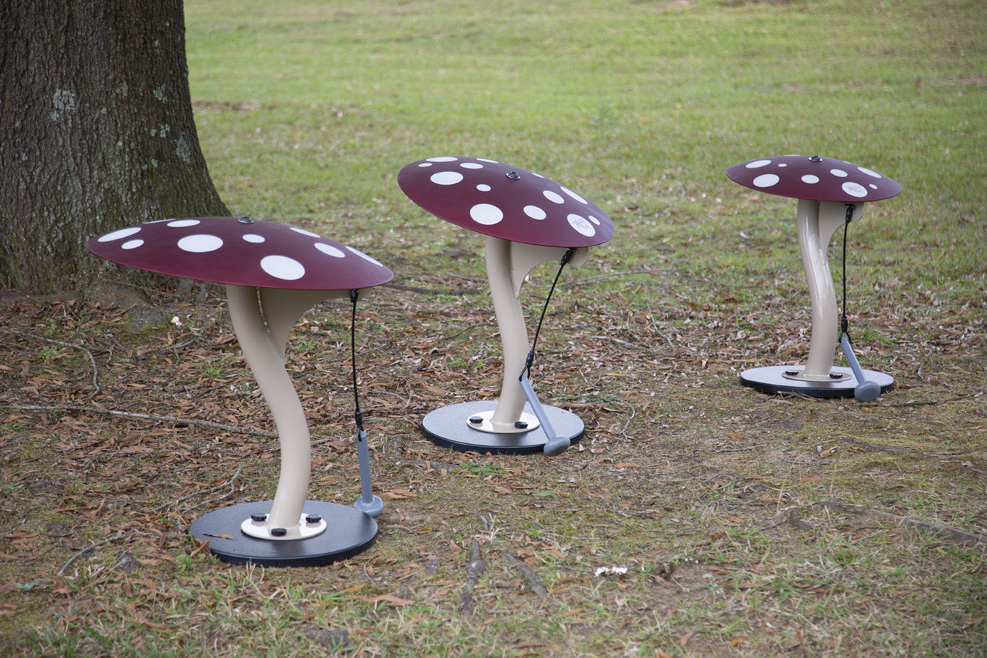 Three red mushroom-shaped outdoor musical instruments with white spots and hanging mallets on grass, producing deep gong sounds.