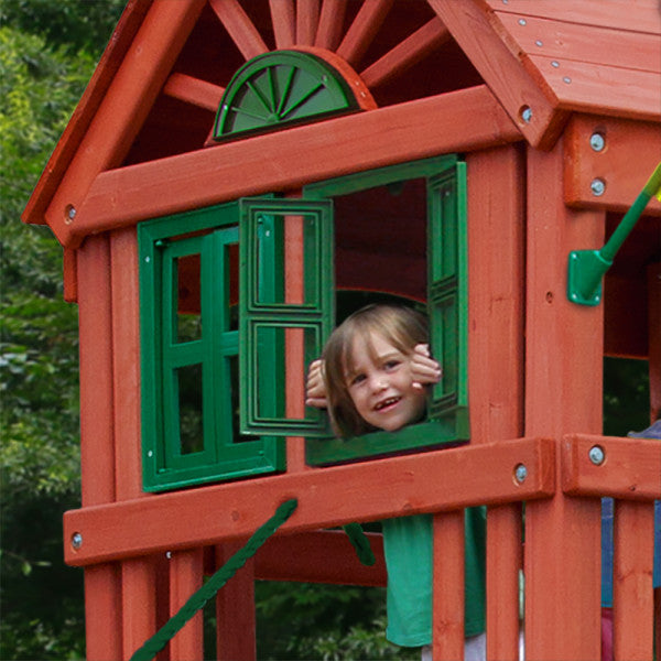 Child peeking through green shuttered window of redwood-finished wooden playset clubhouse with decorative roof details