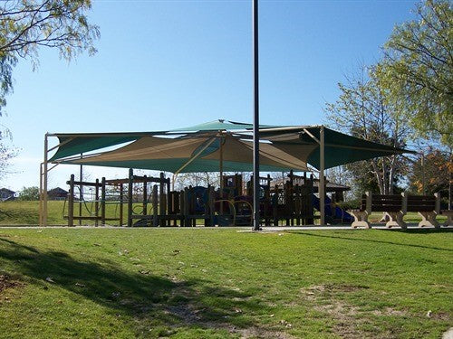 Modern pyramid shade structure with overlapping fabric layers covering playground equipment in a park setting