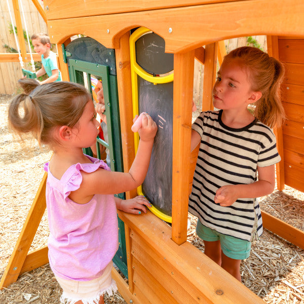 Two children playing on a wooden playset with a working door, double-sided chalkboard, and shaded cafe table with bench.