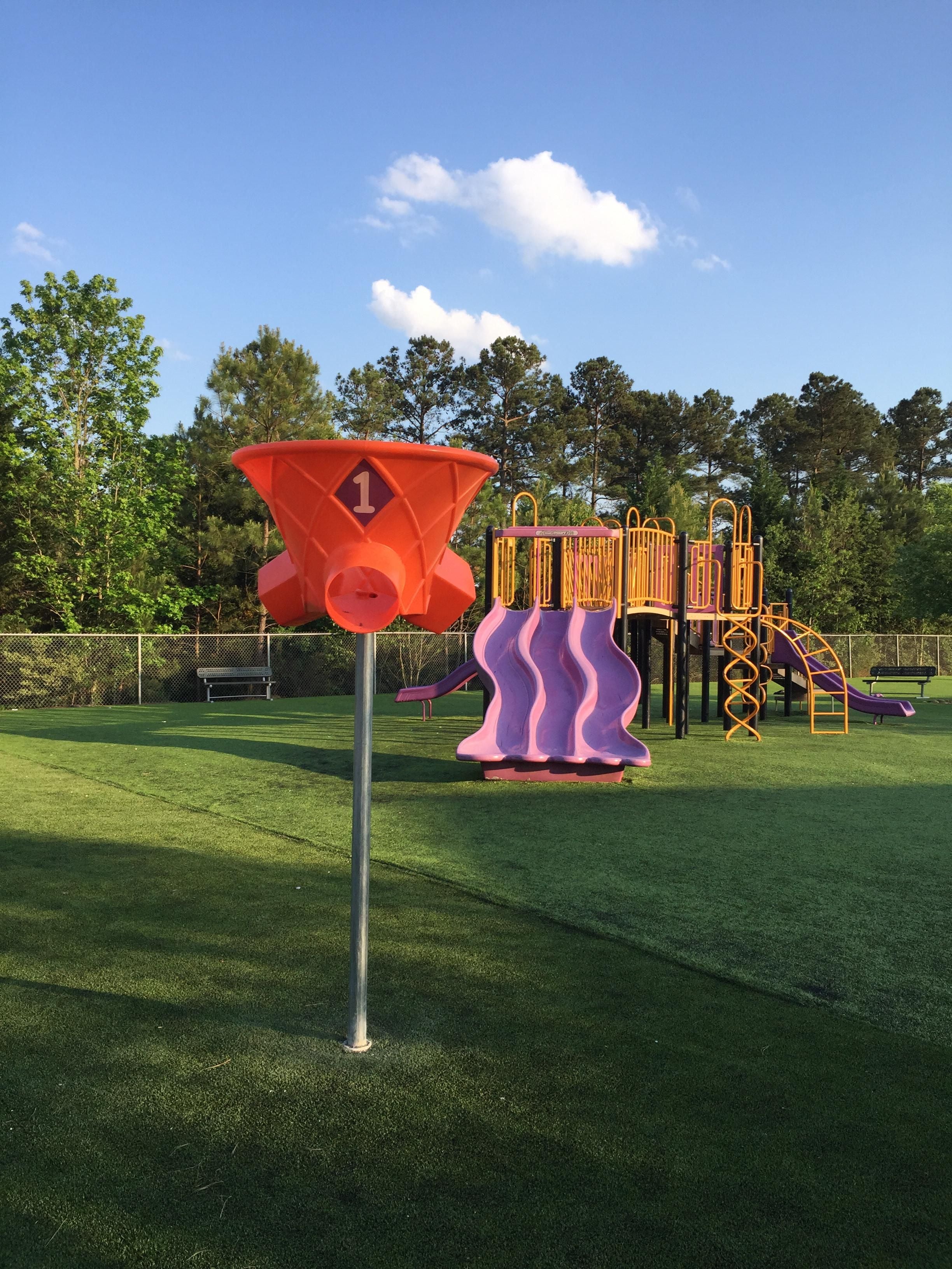 Three-sided orange playground ball game hoop on metal pole with purple slides and climbing structure nearby