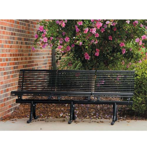 Black metal outdoor bench with individual slatted seats and backrests near flowering shrub and brick wall