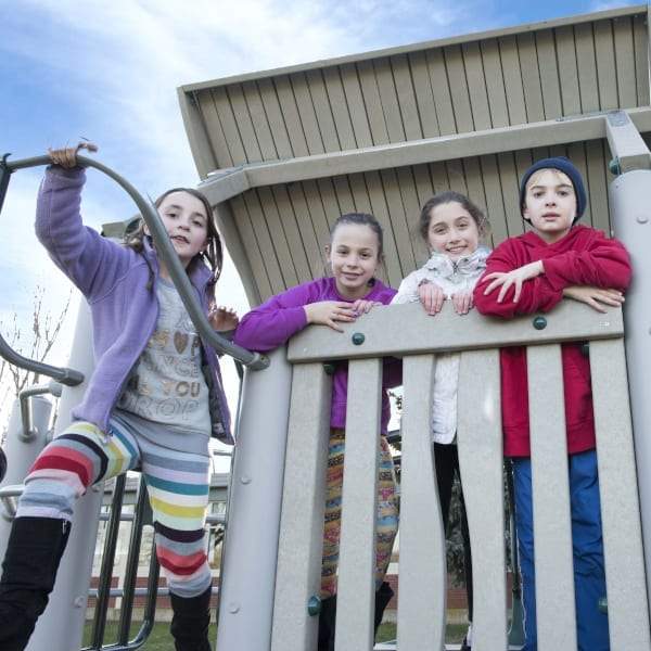 Four children in colorful winter clothes leaning on a wooden railing of a roofed cedar play structure with climbers and pedestals outdoors