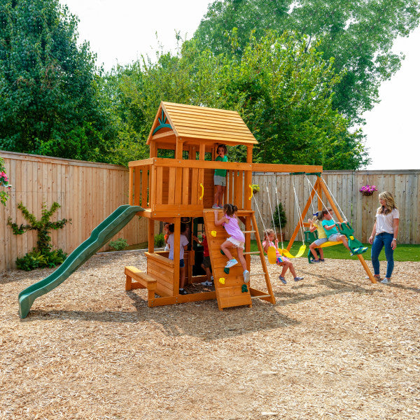 Two-story wooden playset with rock climbing wall, high rail wave slide, two belt swings, glider, and lower clubhouse with play clock and chalkboard.