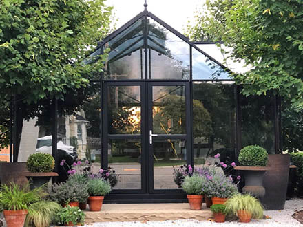 Dark green classic English-style greenhouse with large glass panels, double sliding door, and potted plants around.