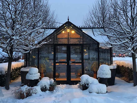 Dark green aluminum-framed greenhouse with tempered glass panels, illuminated interior, surrounded by snow-covered plants and trees.