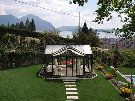 Dark green classic English-style greenhouse with glass panels, roof and louvre windows, set in a landscaped garden with mountain view