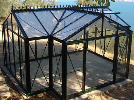 Dark green classic English-style greenhouse with tempered glass panels, decorative roof cresting, and sliding double door
