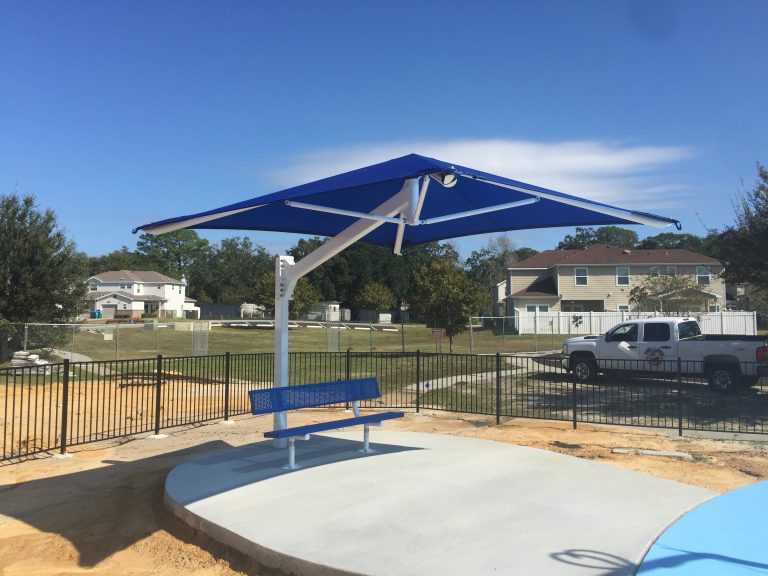 Blue cantilever square shade umbrella with white frame over blue metal bench on concrete pad in outdoor park setting