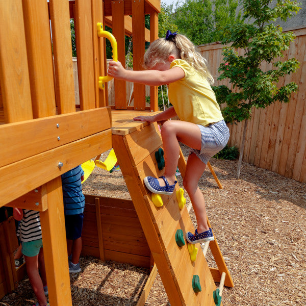 Child climbing wooden rock wall with yellow handholds on backyard playset featuring natural wood finish.