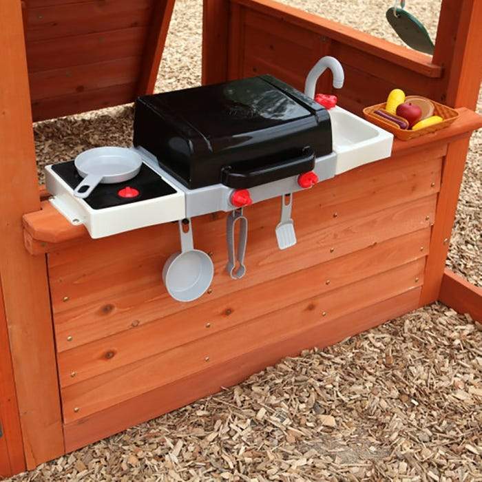 Children's wooden playhouse wall with black grill, white sink, toy utensils, and plastic play food in play kitchen setup