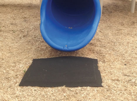 Black rubber playground mat with waffle texture and beveled edges positioned at base of blue plastic slide on mulch surface