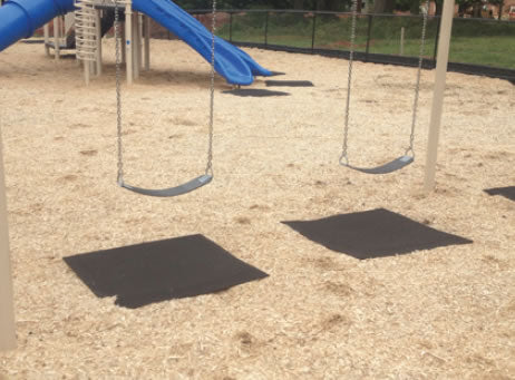 Two black rubber playground mats with beveled edges placed under swings on mulch surfacing to prevent erosion and wear.