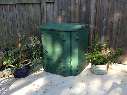 Green insulated compost bin with hinged lid and ventilation panels, placed outdoors on a patio between potted plants