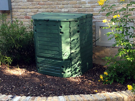 Green insulated compost bin with dual hinged lids, ventilation panels, and large bottom access doors, placed outdoors near plants and brick wall