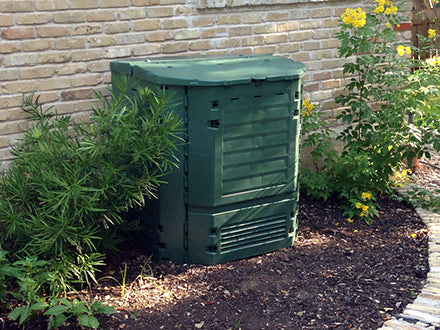 Dark green insulated compost bin with dual hinged access panels, ventilation slots, and a large top lid in a garden setting