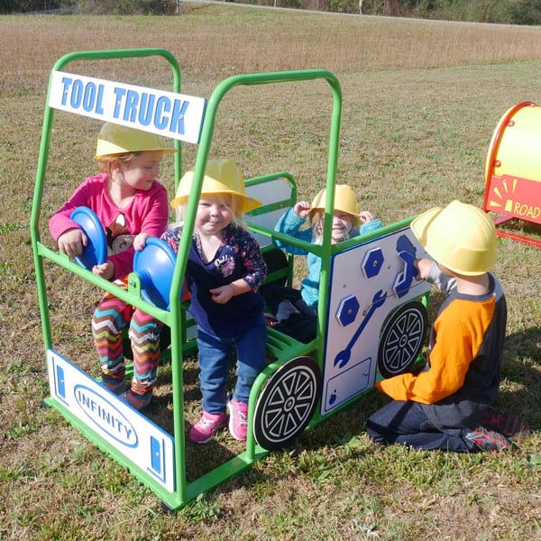 Children wearing yellow helmets playing on a green outdoor tool truck playset with interactive panels and steering wheels