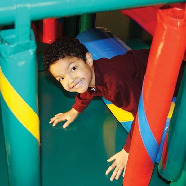 Child crawling through colorful padded tubes in an indoor jungle-themed contained play area with soft foam flooring and protective netting.