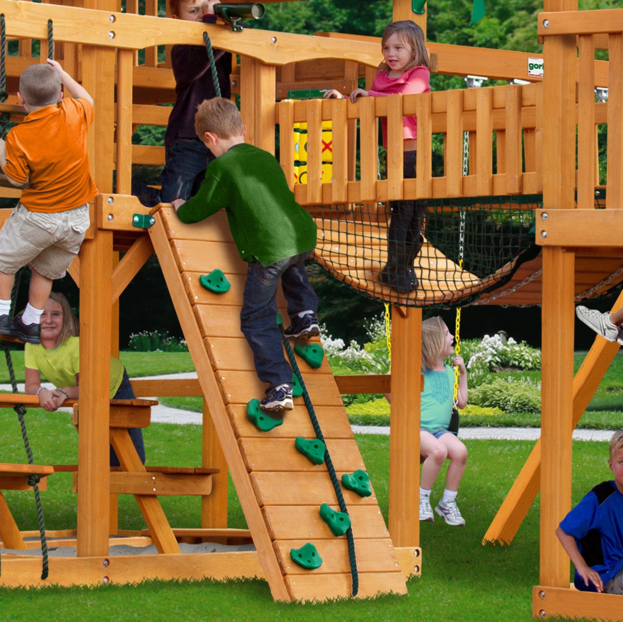 Cedar wooden playset with climbing wall, rope bridge, swings, and children playing on grass outdoors