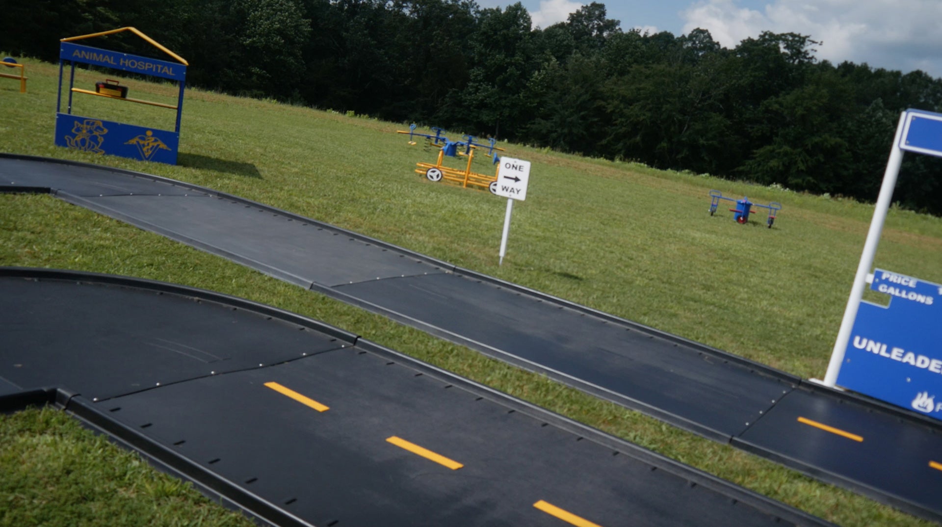 Curved black trike path with yellow lane markings, miniature road signs, and playground structures on grass