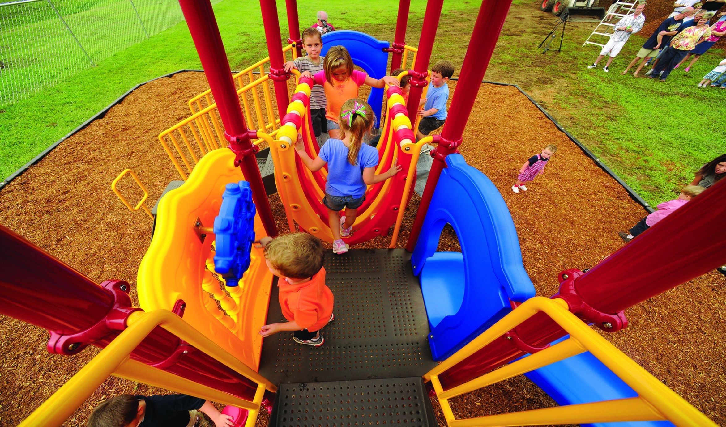 Children playing on colorful playground with slides, climbers, interactive panels, and wood chip ground under large shade structure