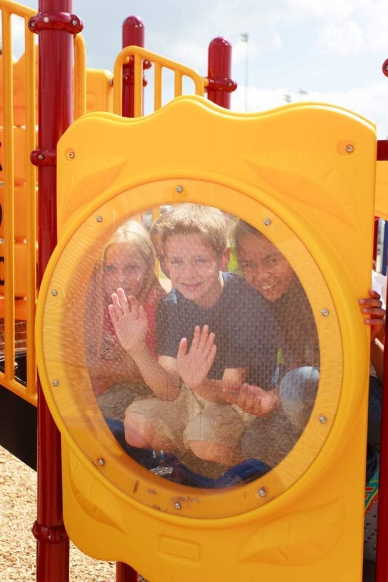 Three children smiling behind a circular yellow play panel on a colorful playground structure with climbing elements and slides.