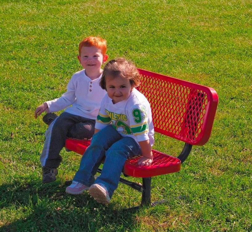 Red portable preschool bench with diamond-patterned metal seat and backrest, two young children seated outdoors on grass