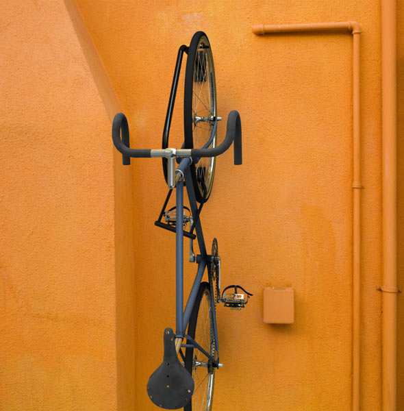 Vertical bike mounted on black wall rack against textured orange wall with visible pipes and utility box