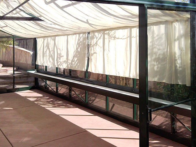 Long black seed tray shelf with corrugated plastic bottom inside a Royal Victorian greenhouse with translucent curtains