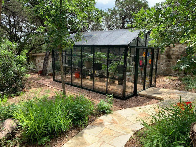 Black-framed Victorian-style glass greenhouse with pitched roof, surrounded by lush garden plants and stone pathway.