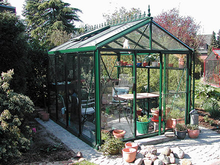 Dark green Victorian-style glass greenhouse with pitched roof, multiple roof windows, and potted plants inside and outside.