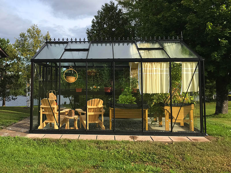 Black-framed glass Victorian greenhouse with roof windows, wooden chairs, hanging basket, and raised garden beds with plants inside