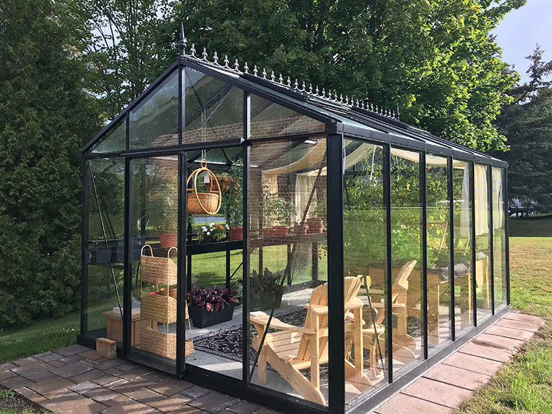 Black-framed Victorian-style glass greenhouse with decorative roof finials, interior plants, wooden chairs, and hanging baskets in garden setting