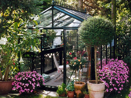 Black-framed glass Victorian greenhouse surrounded by lush greenery and vibrant pink flowers in a garden setting