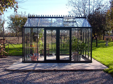 Classic black-framed glass greenhouse with pitched roof, double doors, and potted plants inside on concrete base