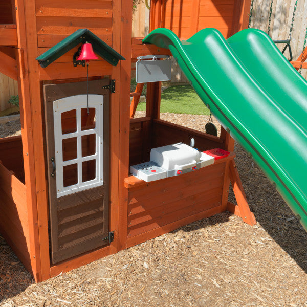 Wooden playset corner with white magnetic door, red bell, built-in BBQ kitchen, and green double wave slide