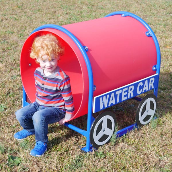 Child inside red and blue cylindrical play structure with wheel designs on grass, part of construction-themed playground set